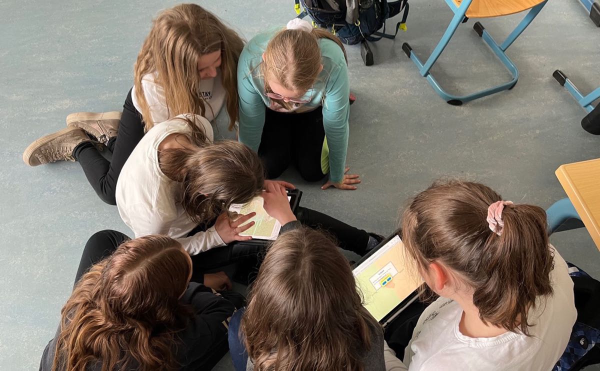 Foto, auf dem sechs Schülerinnen zu sehen sind, die auf dem Boden eines Klassenzimmers sitzen und gemeinsam an Tablets arbeiten.