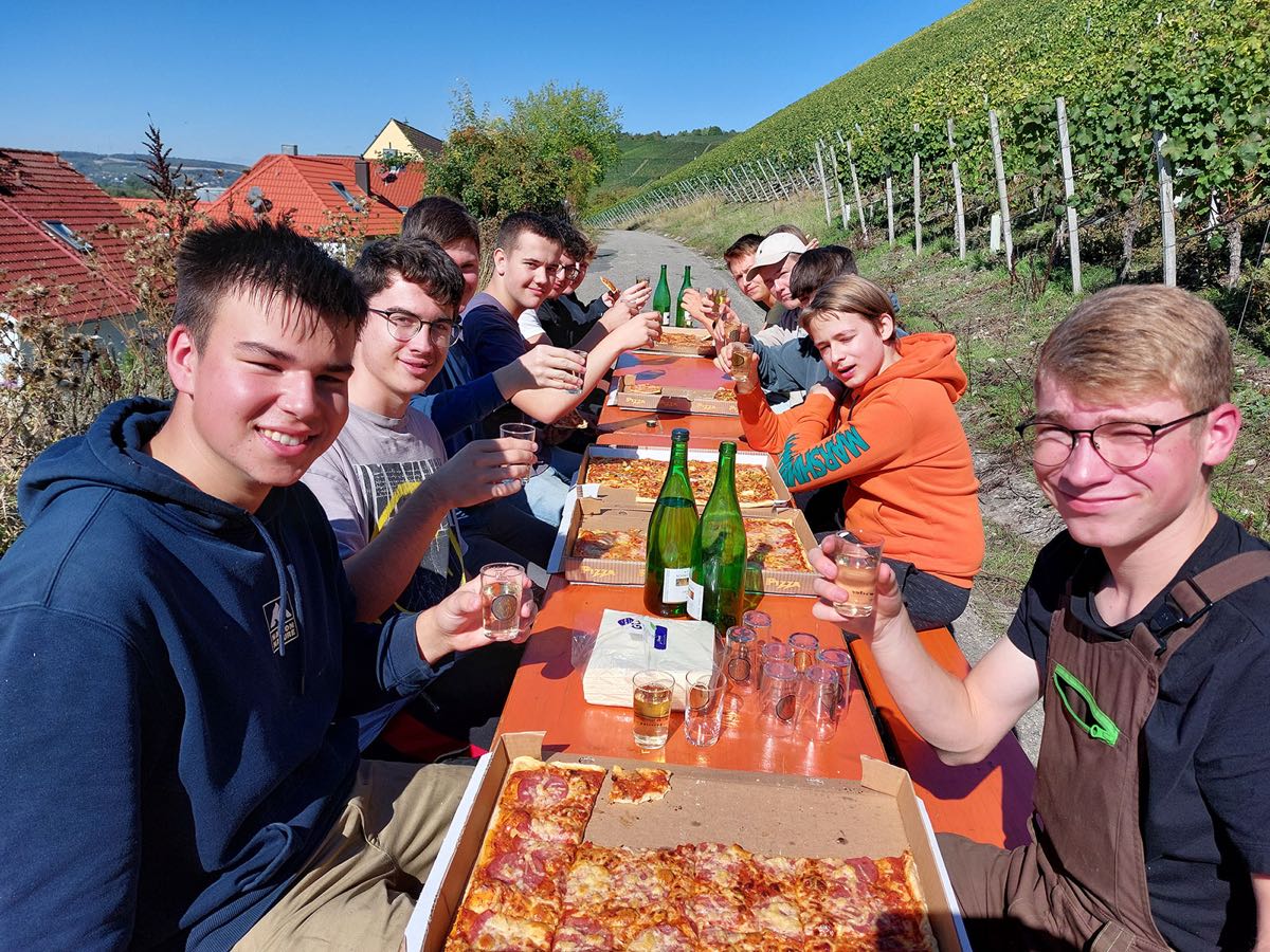 Zu sehen sind Schülerinnen und Schüler, die im Freien an einer Bierbank sitzen und Pizza essen. Im Hintergrund ist ein Weinberg zu sehen.