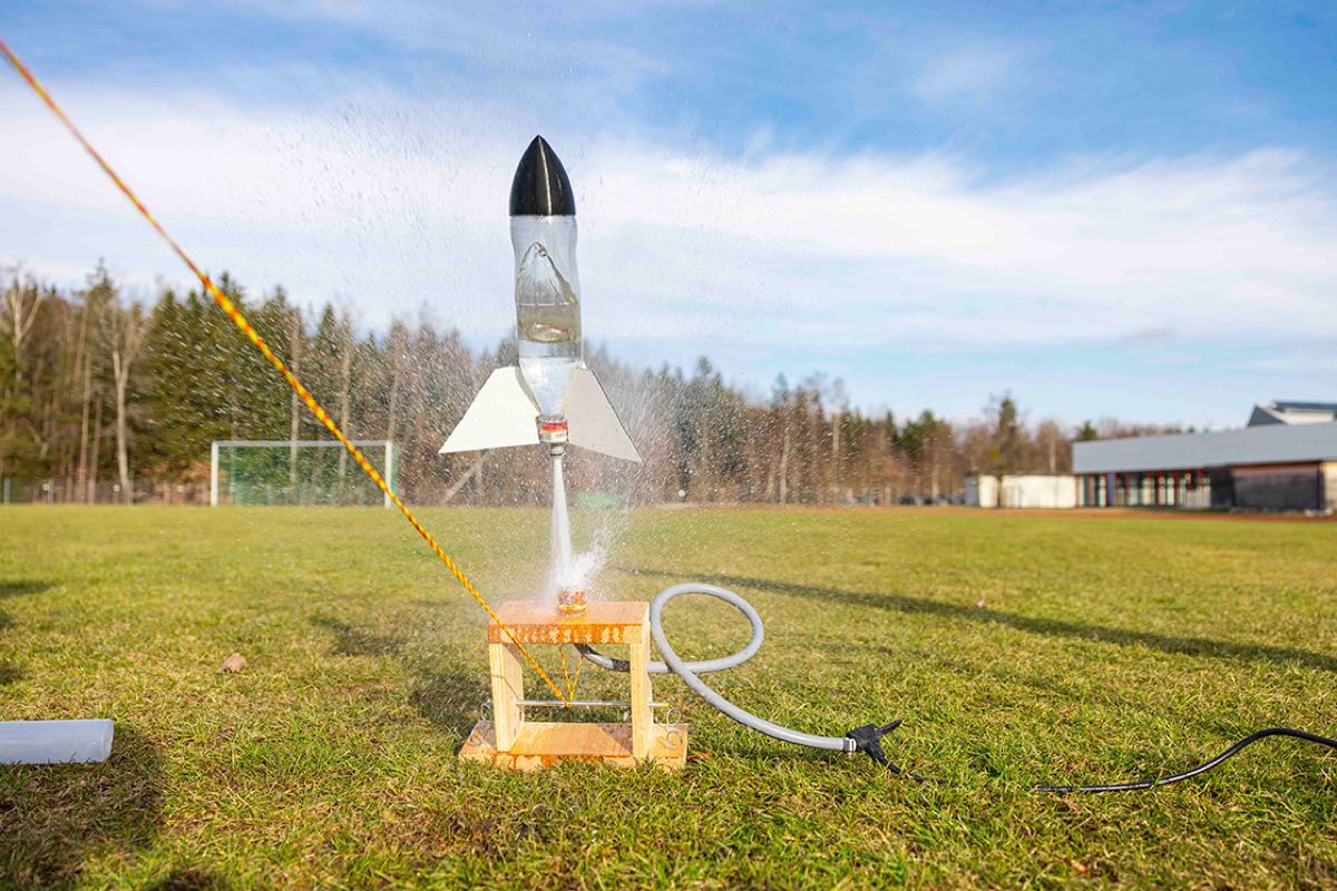 Zu sehen ist eine selbstgebaute Rakete aus einer Wasserflasche und einem Gartenschlauch auf einer großen Wiese. Im Hintergrund sind ein Fußballtor und ein flaches Gebäude zu sehen.